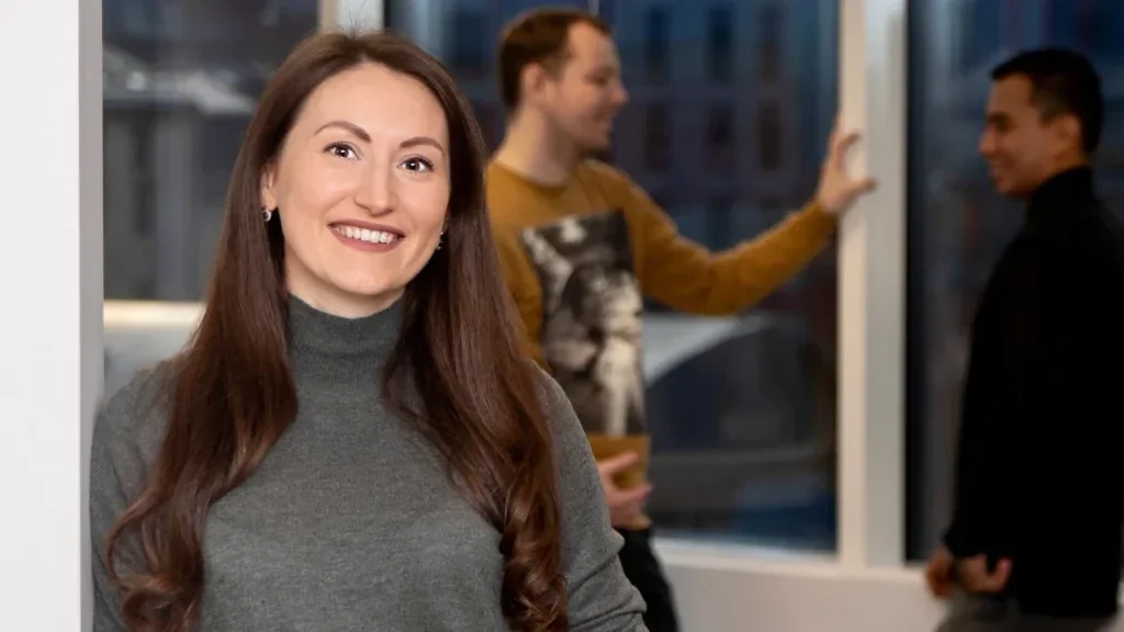 Smiling woman holding a coffee mug in an office, with two men talking by the window behind her