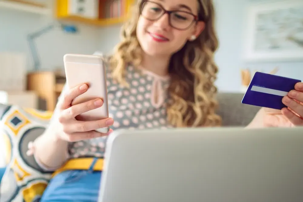 Woman on a sofa shopping online with a phone, a laptop and a credit card